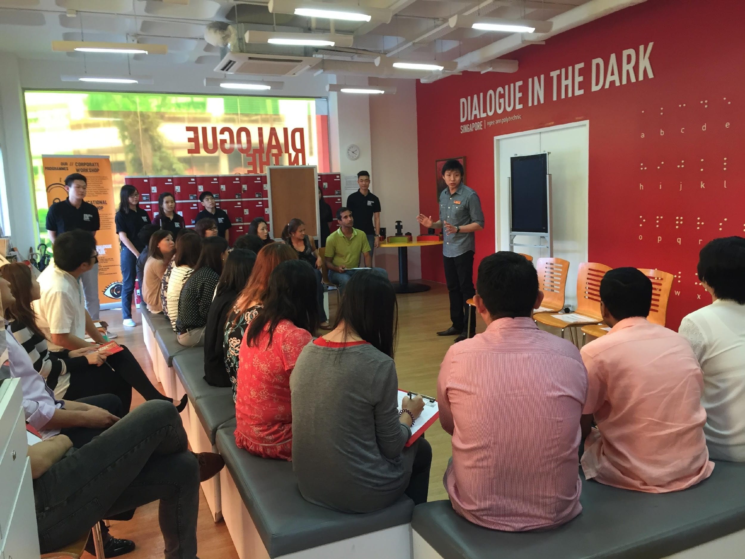 A presenter speaks to a seated audience inside the Dialogue in the Dark Singapore venue with braille on the red wall.